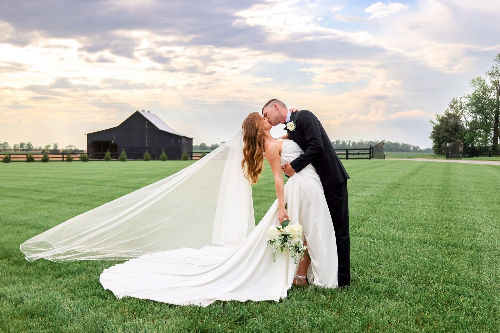 Bride and groom kissing in the open pasture with the black barn and evening sky behind them